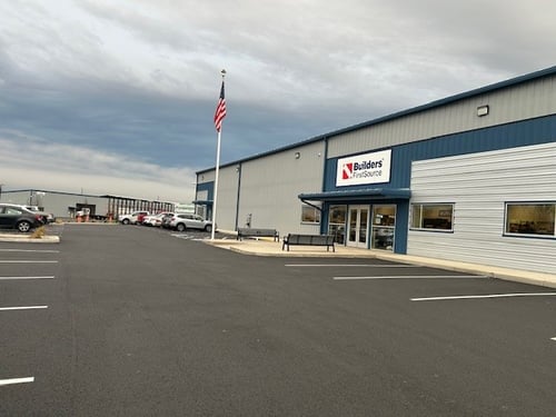 Front entrance of the Builders FirstSource location in Woodland, WA, featuring a freshly paved parking lot, American flagpole, and glass entry doors on a modern metal building with blue trim.