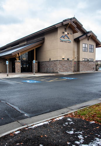 An exterior shot of the "Idaho First Bank" building with its distinctive lodge architecture, including a prominent roof and stone facade. The parking lot in the foreground has marked handicap parking spaces, and there's some sparse foliage and a bit of snow on the ground.