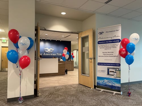 Photo of open double doors leading to the reception area of the First American Title office in Anchorage, AK. Red, white and blue balloons are on both sides of the doors with a vertical banner on the right side explaining the services offered.
