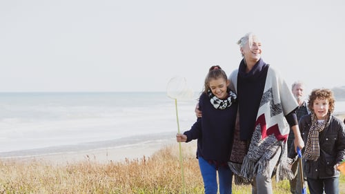 A joyful family strolls along a grassy beach path, with the ocean in the background. A young girl holds a fishing net, smiling alongside an older woman.