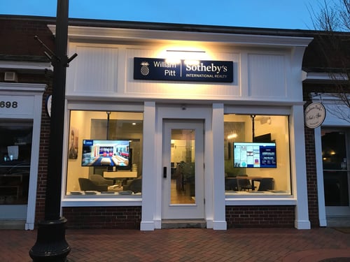 White storefront with brick base and large display windows housing William Pitt Sotheby's International Realty in Madison, Connecticut, photographed at dusk with illuminated interior showing digital property displays.