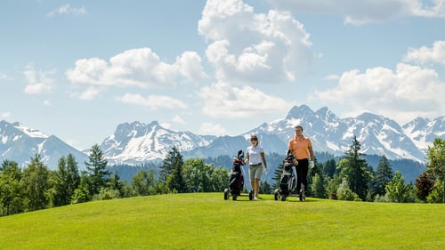 Zwei Golfer laufen mit Golfbags auf dem Green des Golfplatzes Sonnenalp – im Hintergrund sind die schneebedeckten Allgäuer Berge.