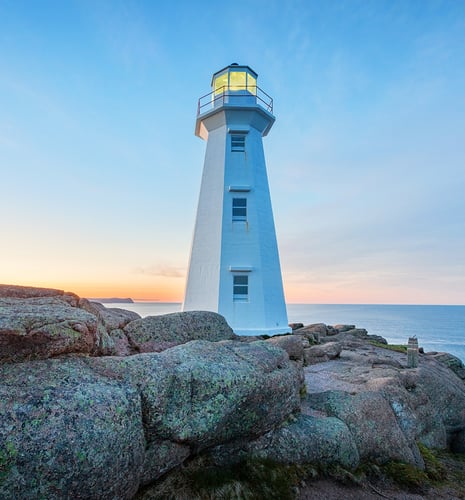 White lighthouse on rocky coast at sunset, with a glowing lantern room. The sky is a gradient of soft orange to blue, conveying calmness.