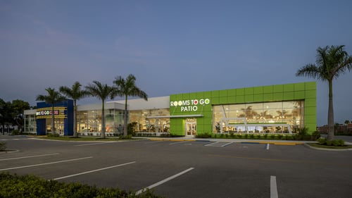 The Rooms To Go Kids and Patio showroom exterior featuring vibrant green and blue facades, large glass windows, and palm trees under a twilight sky.