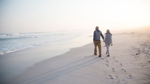 An elderly couple walks hand in hand along a serene beach at sunrise, casting elongated shadows on the sand. Waves gently lap the shore, creating a peaceful atmosphere.