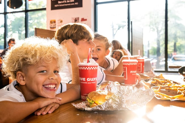 A child smiles while enjoying a Five Guys burger with friends at a Five Guys restaurant.