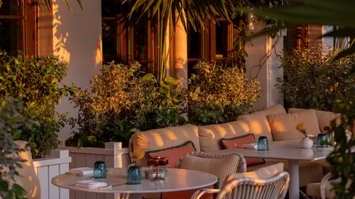 Outdoor restaurant seating area with cushioned chairs, round tables, candle holders and surrounding lush plants in warm sunset lighting