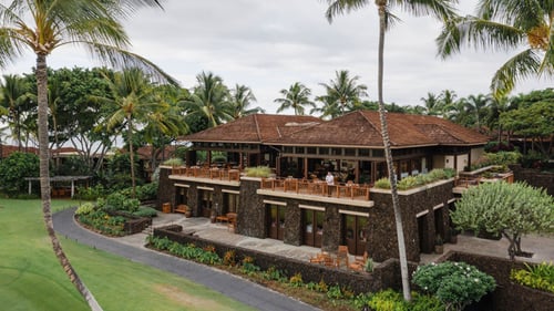 Tropical restaurant with stone walls, wooden balconies and an open-air dining area, surrounded by lush greenery and palm trees