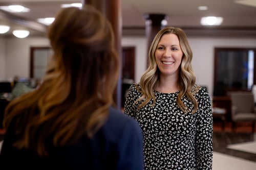 A smiling Bryant Bank employee in a leopard-print blouse assists a customer at a polished wood teller desk. The customer, viewed from the back, is wearing a navy blazer and holding documents during the interaction.