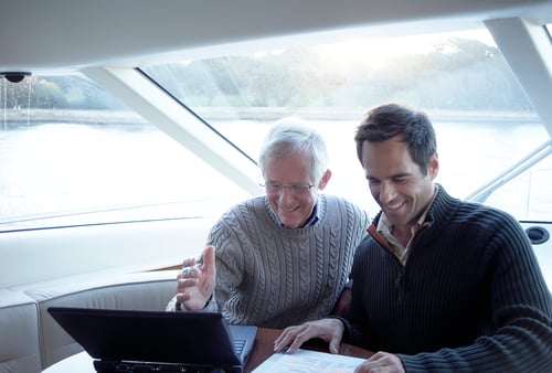 Two men smiling and engaged in using a laptop on a boat. One gestures at the screen, wearing a gray sweater, while the other wears a dark sweater. Sunlight streams through large windows, and a serene water view is visible.