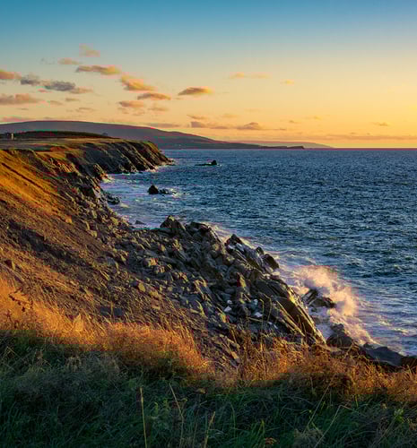 Rocky coastline at sunset with waves crashing against the shore. The golden light enhances the rugged cliffs and calm, expansive sea, creating a serene atmosphere.