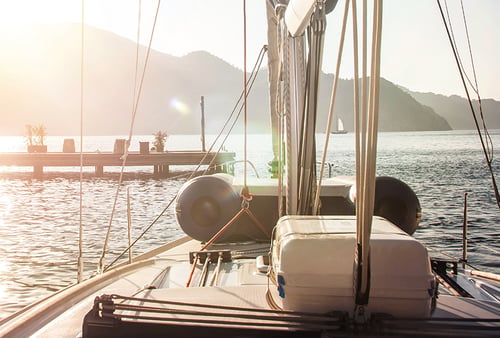 Sunlit sailboat deck facing calm waters, with distant mountains and a sailing boat on the horizon, conveys a serene and peaceful atmosphere.
