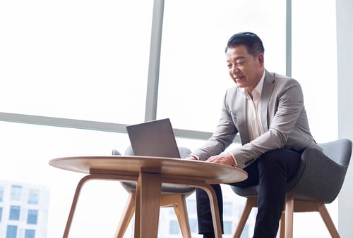 A man in a gray suit sits on a gray chair, smiling and using a laptop on a wooden table. Bright, modern office with large windows in the background.