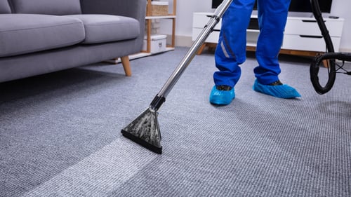 An eye-level, medium shot shows a professional cleaner in blue overalls and blue shoe covers using a silver and black steam cleaner on a grey textured carpet in a living room. The cleaner is moving the steam cleaner from the bottom left to the top right of the frame, leaving a bright white streak on the carpet. The cleaner's legs and the bottom of their blue overalls are visible, as well as the black hose of the steam cleaner. In the background, there is a grey sofa on the left and a white media console with drawers on the right. The floor is covered in a grey textured carpet. The lighting is bright and even, suggesting a professional cleaning service.