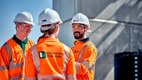 Three employees in safety glasses, white hard hats, and orange reflective jackets with Heidelberg Materials branding converse at an industrial site.