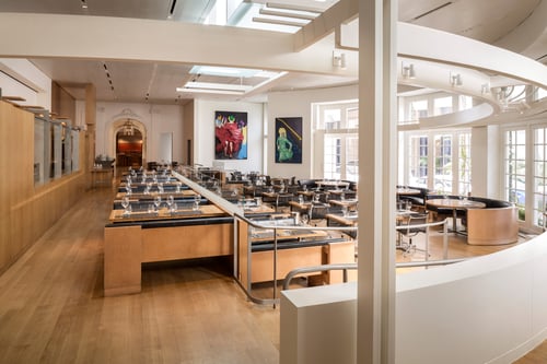A modern restaurant dining area with sleek wooden tables and glassware set for service. Large windows allow natural light, and vibrant artwork decorates the walls.