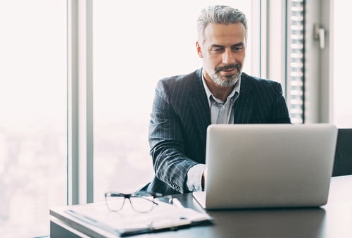 Businessman in a suit working on a laptop at an office desk with eyeglasses and documents. He looks focused, with city skyline visible outside.