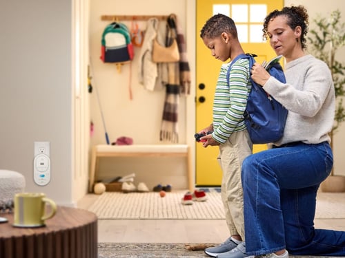 A mom helps her son put his backpack on, and a co alarm is plugged into the background.