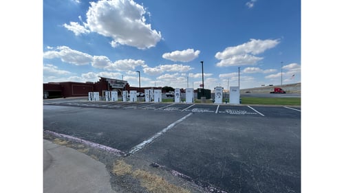 Several EVgo fast charging units installed in marked parking spaces at a public charging station, with EVgo logos on the chargers, protective bollards around each unit. CCS chargers featured in the image. HomeZone furniture store is visible in the background.