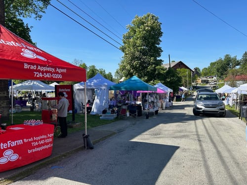 A vibrant street fair scene with colorful tents, a State Farm booth, and visitors browsing merchandise under clear blue skies.