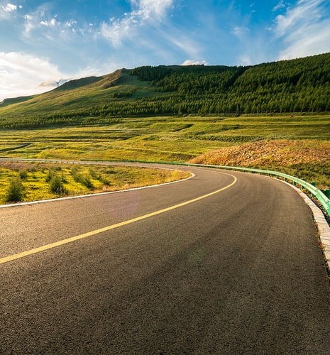Curving road through lush green hills under a clear blue sky with scattered clouds; wind turbine visible on distant hill, conveying serenity.