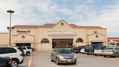 Gold car driving between rows of parked cars in front of Tom Thumb shopping center