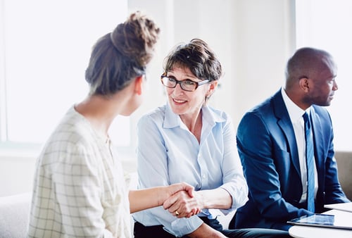 Two women sit in an office setting, warmly shaking hands and smiling, suggesting a positive interaction. A man in a suit looks away thoughtfully.