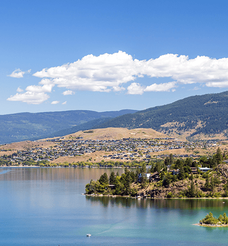 Scenic view of a calm lake reflecting a partly cloudy sky. Hills with scattered houses line the shoreline, backed by lush green mountains. Bright, peaceful atmosphere.