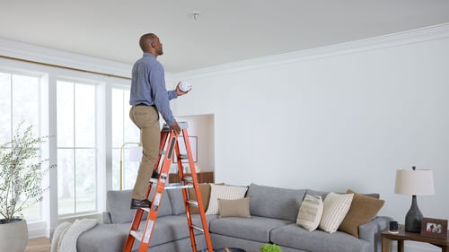 A man on a ladder is installing a new smoke alarm.