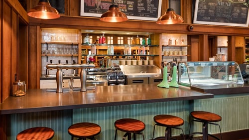 Cozy café bar with wooden stools, a counter with drink taps, hanging copper lights and a menu on chalkboards above the shelves stocked with glasses and bottles