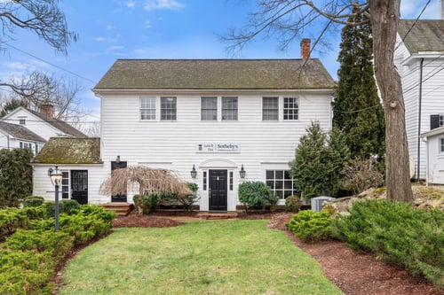 White colonial-style building with black door and arched entrance housing Julia B Fee Sotheby's International Realty in Bedford, New York featuring landscaped lawn and evergreen plantings.