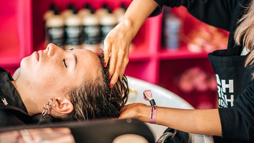 Person with their head resting in a shampooing basin whilst a member of staff applies product to their hair. Colourful product display in background.
