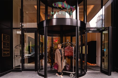 Stylish woman exiting Marcus restaurant through a revolving door, carrying shopping bags and dressed in a chic coat, with the store name 'Marcus' displayed above.