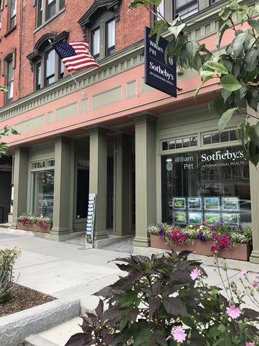 Pink and brick building with cream-colored columns and storefront housing William Pitt Sotheby's International Realty in Great Barrington, Massachusetts, featuring American flag and colorful flower planters.