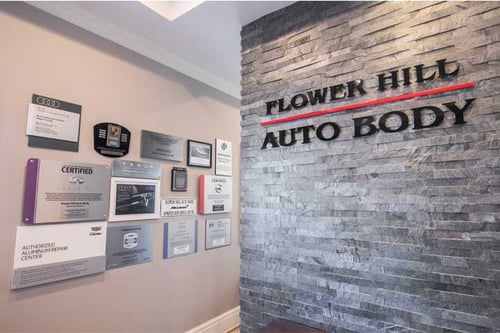 The image shows an interior wall of “Flower Hill Auto Body,” featuring the company’s name in large black letters with a red line under “Flower Hill” on a textured gray stone wall. Adjacent to the company name, a collection of certification plaques and awards is displayed on a beige wall, representing the shop’s expertise and recognized standards in auto body repair.
