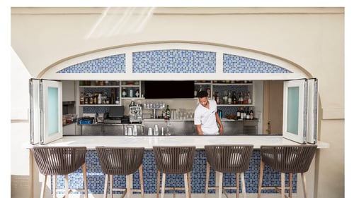 A bartender in a white shirt preparing a poolside bar with blue mosaic tiles, surrounded by four high-back wooden chairs. Bottles and glasses are visible on shelves behind him.