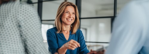 Une femme souriante vêtue d'un chemisier bleu converse avec deux personnes de l'autre côté d'une table, dégageant une atmosphère chaleureuse et professionnelle dans un bureau moderne.