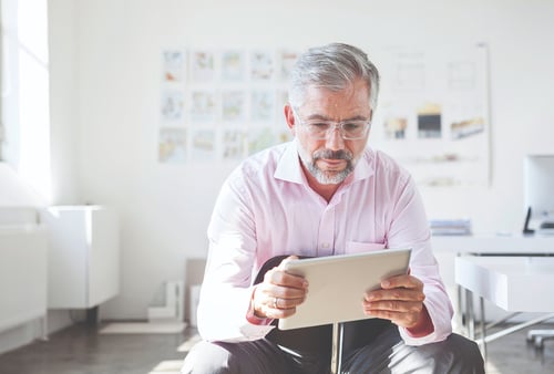 Elderly man with glasses in a light pink shirt sits in a bright office, reading a tablet. White walls with blurred artwork create a peaceful atmosphere.