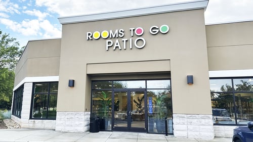 Exterior of a Rooms To Go Patio store with large glass windows, tan facade, and clear signage above the entrance on a partly cloudy day.