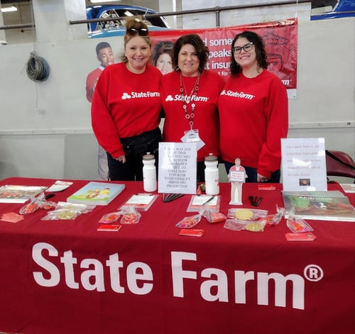 Agent Debbi and two team members standing and smiling together wearing red State Farm shirts behind red State Farm table