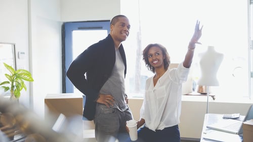 Two people in a bright space discuss ideas. A woman, smiling, gestures upward with enthusiasm, holding a coffee cup. A man stands beside her, listening intently.