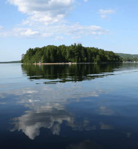 A wooded island reflected in the calm, mirror-like surface of a lake, with fluffy white clouds visible in the sky and the water below.