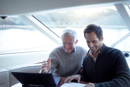 Two men smiling and engaged in using a laptop on a boat. One gestures at the screen, wearing a gray sweater, while the other wears a dark sweater. Sunlight streams through large windows, and a serene water view is visible.