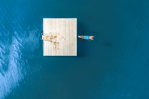 Overhead view of a person in blue shorts floating calmly next to a wooden dock in deep blue water, evoking a sense of tranquility and solitude.