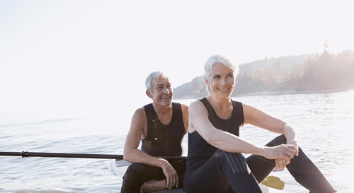 A smiling older couple in wetsuits sit on paddleboards on a calm lake, bathed in sunlight. The scene conveys tranquility and joy.