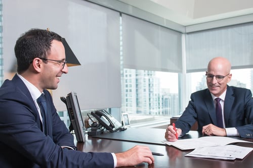 Two male professionals in navy suits sitting across from each other at a dark wooden desk in a modern office with floor-to-ceiling windows overlooking a city skyline. They appear to be in a meeting or consultation, with documents on the desk between them. The office features contemporary furnishings including a desk lamp and office equipment, creating a polished, corporate atmosphere.