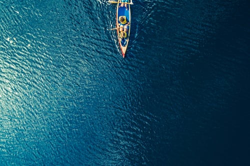Aerial view of a small, colorful fishing boat with two people, surrounded by vast, rippling blue water. The scene conveys peacefulness and solitude.