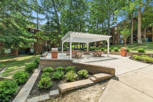 A patio with a white pergola and a fire pit surrounded by green bushes.