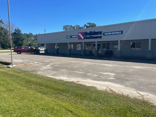 A storefront for "Builders FirstSource" is shown, featuring a white metal facade with signage that includes the store name and the phrases "Lumber • Hardware" and "Building Materials." The building has multiple glass windows and a covered entrance supported by stone columns. A red pickup truck is parked on the left side of the image, with a grassy area and flagpole in the foreground. The sky is clear and blue, indicating a sunny day. The parking lot in front of the store is mostly empty.