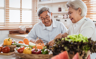 Elderly couple in a bright kitchen smiling as they chop vegetables. Fresh produce like peppers and greens surround them, creating a warm, joyful atmosphere.